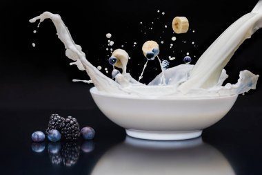 Blackberries and banana dropping into white bowl with milk on black background clipart