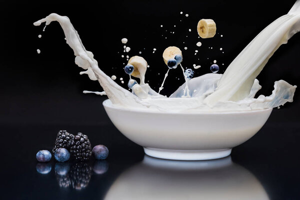Blackberries and banana dropping into white bowl with milk on black background