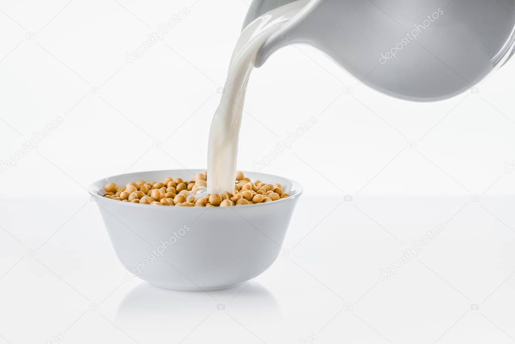 Pouring milk from jug in bowl with soybeans on white background