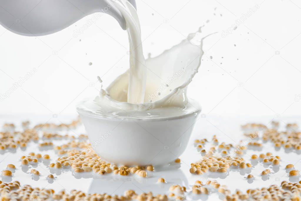 Milk splashing from jug in bowl with soybeans on white background