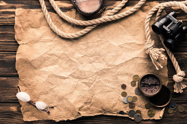 top view of blank crumpled paper with compass, binoculars and rope on rustic wooden surface