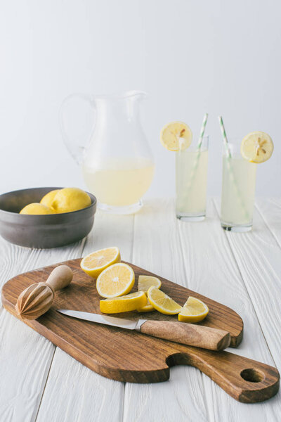 close up view of lemons, knife, wooden pestle on cutting board and lemonade in glasses on white wooden tabletop on grey backdrop