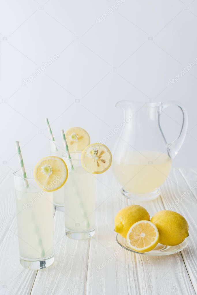 close up view of lemonade in jug and glasses with straws on white wooden tabletop on grey background