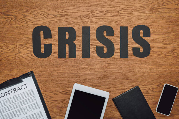 top view of word crisis near smartphone, digital tablet, notebook and clipboard with contract on wooden desk