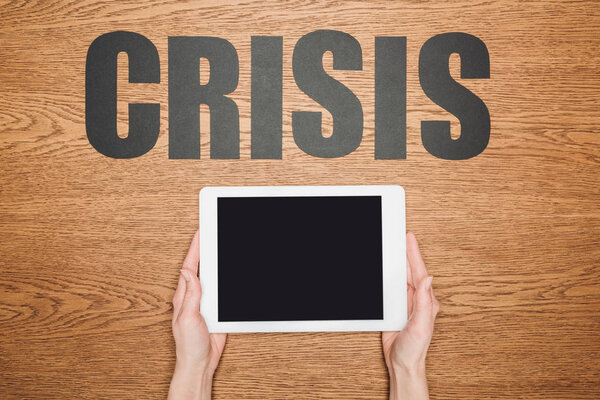 cropped view of woman holding digital tablet with blank screen near black paper cut word crisis on wooden desk