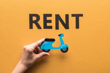 Kırpılmış görüntüde elinde kağıt kesilmiş scooter olan kadın turuncu arka planda kiralık harflerin yanında.