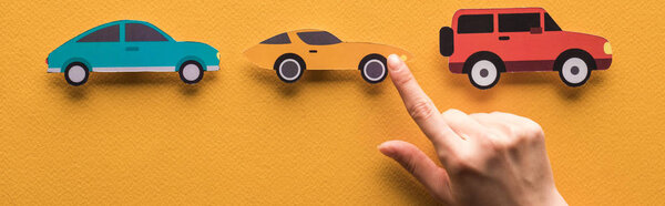 обрезанный вид женщины, указывающей пальцем на разрезанные бумагой машины на оранжевом фоне, панорамный снимок
