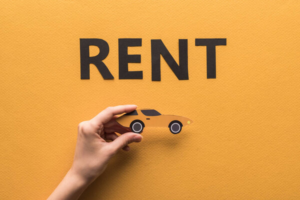 cropped view of woman holding paper cut sports car near rent lettering on orange background
