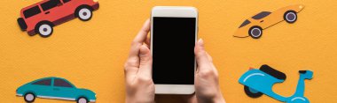 Beyaz ekranlı akıllı telefonu olan, turuncu arka planda kağıt arabaların yanında panoramik çekim yapan kadın görüntüsü.