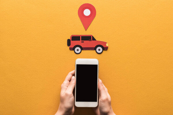 cropped view of woman holding smartphone with blank screen near paper cut car, location mark on orange background
