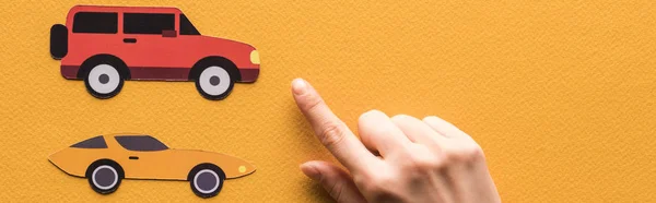 cropped view of woman pointing with finger at paper cars on orange background, panoramic shot