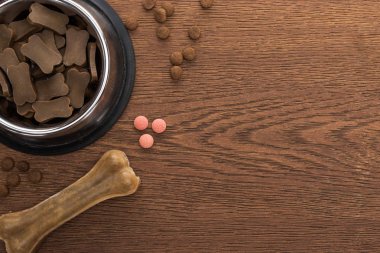 Gümüş kasedeki kuru hayvan mamasının üst görüntüsü kemik ve ahşap masadaki pembe vitaminler.