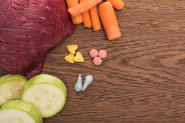 Tahta masadaki vitaminlerin yanında çiğ et, kabak ve havuç manzarası.