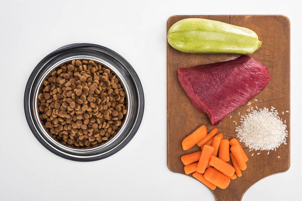 top view of dry pet food and raw meat, rice, carrot and zucchini on wooden cutting board isolated on white