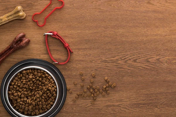 top view of dry pet food in silver bowl near bones, collar and pet toy on wooden table