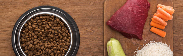 вид сухой корм для домашних животных в серебряной миске рядом с сырым мясом, цуккини, рисом и морковью на деревянном столе, панорамный снимок
