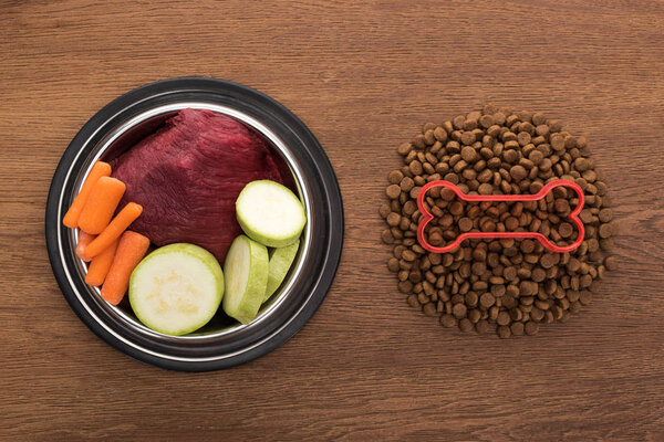 top view of dry pet food with bone near raw vegetables and meat in bowl on wooden table