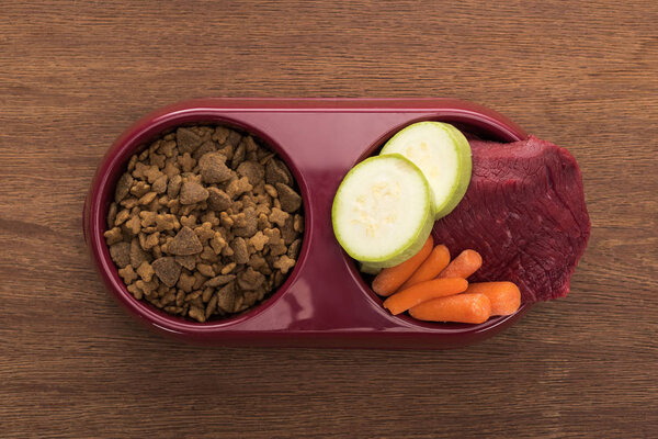 top view of dry pet food and raw meat with vegetables in bowl on wooden table