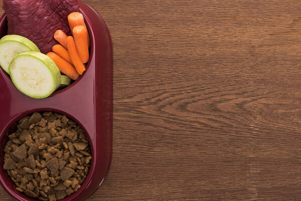 top view of dry pet food and raw meat with vegetables in bowl on wooden table with copy space