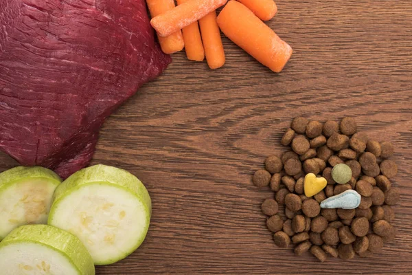 top view of raw meet, zucchini and carrot near vitamins on dry pet food on wooden table