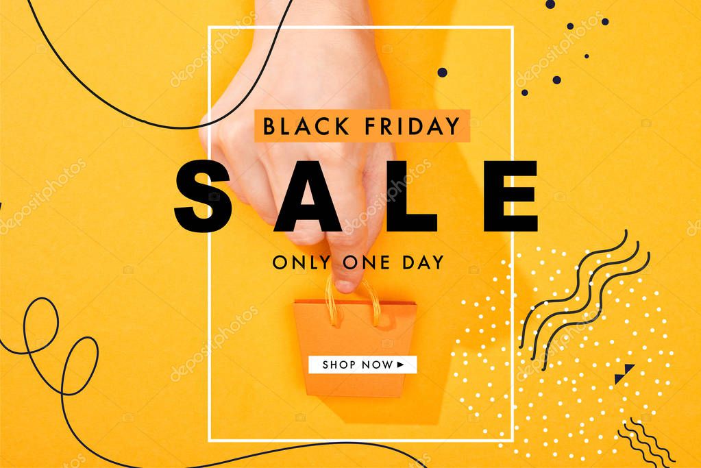 Cropped view of hand holding small shopping bag on bright orange background with black Friday sale illustration