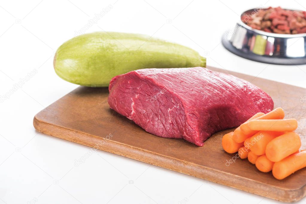 selective focus of dry pet food in bowl near raw meat, carrot and zucchini on wooden cutting board isolated on white