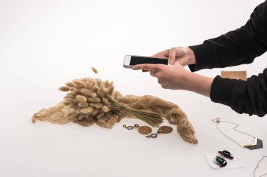 cropped view of photographer taking pictures of composition with flora and jewelry on smartphone on white clipart