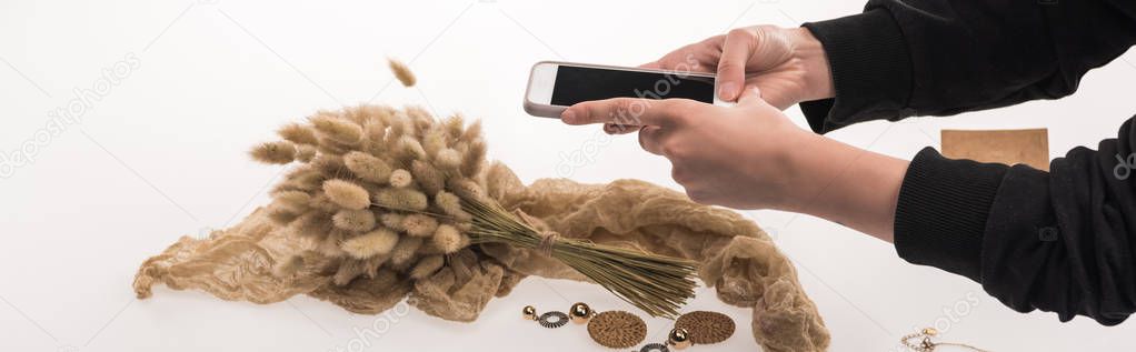 partial view of commercial photographer taking pictures of composition with flora and jewelry on smartphone on white, panoramic shot