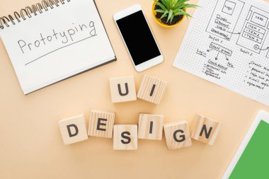 Tahta blokların yakınındaki cihazların üst görünümü ui tasarım harfleri, web sitesi tasarım şablonu, prototip harfleri ve bej arka planda yeşil bitki içeren defter