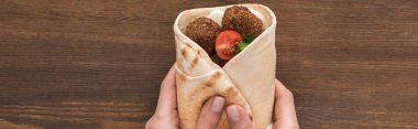 cropped view of woman holding falafel with vegetables and sauce in pita on wooden table, panoramic shot clipart