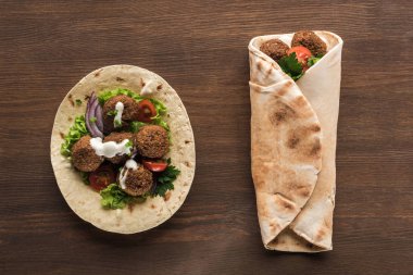 top view of falafel with vegetables and sauce wrapped and unwrapped in pita on wooden table clipart