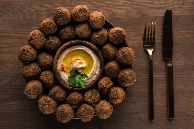 Tahta masadaki çatal bıçak takımının yanında humus kaplı falafel toplarının üst görüntüsü.
