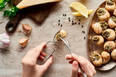 cropped view of woman eating escargots with tweezers near lemon slices, Parmesan, garlic, parsley, black peppercorn on stone background clipart
