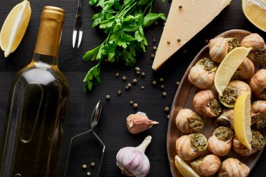 Lezzetli, pişmiş salyangozlar, siyah ahşap masa üzerinde limon dilimleri, baharatlar, parmesan ve beyaz şarap.