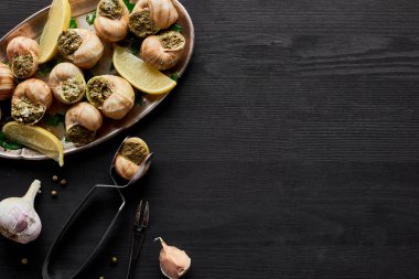 top view of delicious cooked escargots with lemon and tweezers on black wooden table clipart