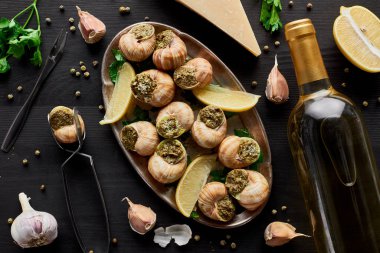 Lezzetli, pişmiş salyangozlar, siyah ahşap masa üzerinde limon dilimleri, baharatlar, parmesan ve beyaz şarap.
