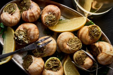 top view of delicious cooked escargots with lemon slices and cutlery on plate on black wooden table clipart