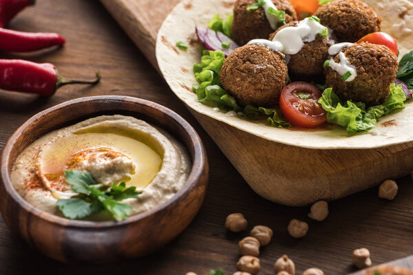 close up view of falafel with vegetables and sauce on pita near hummus on wooden table