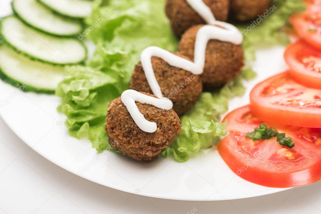 close up view of falafel with sauce on plate with sliced vegetables on white background