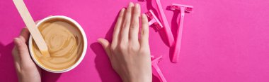 cropped view of woman moving away disposable razors and taking depilation wax in cup with stick on pink background, panoramic shot clipart