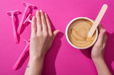 cropped view of woman moving away disposable razors and taking depilation wax in cup with stick on pink background clipart