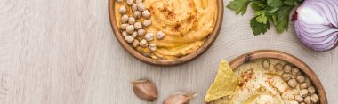 top view of delicious hummus with chickpeas, nacho in bowls near spices and parsley on beige wooden table, panoramic shot clipart