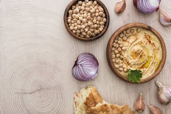 top view of delicious hummus with chickpeas in bowl near fresh baked pita, garlic and red onion on beige wooden table