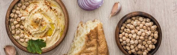 top view of delicious hummus with chickpeas in bowl near fresh baked pita, garlic and red onion on beige wooden table, panoramic shot