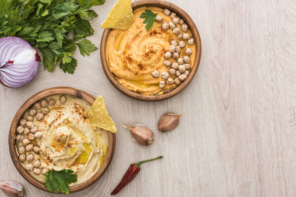 top view of delicious hummus with chickpeas, nachos in bowls near spices and parsley on beige wooden table
