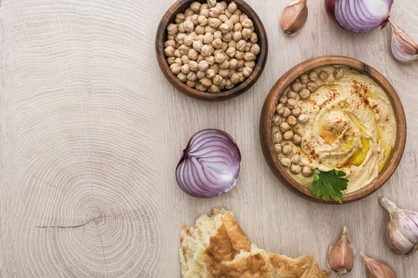 Top view of delicious hummus with chickpeas in bowl near fresh baked pita, garlic and red onion on beige wooden table — Stock Image Top View Delicious Hummus Chickpeas Bowl Fresh Baked Pita Garlic Stock Picture