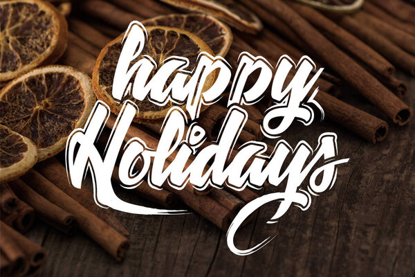 close up view of dried orange slices and cinnamon on wooden background with happy holidays illustration