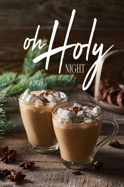 selective focus of cacao with marshmallow and cacao powder in mugs near pine branches, cinnamon and anise on wooden table with oh holy night illustration