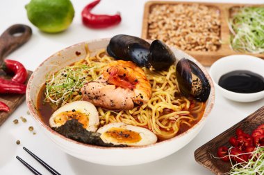 Taze malzemelerin yanında baharatlı deniz ürünleri ramen ve beyaz yüzeyde yemek çubukları.