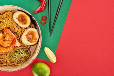 Taze malzemelerin yanında baharatlı deniz mahsulü ramen ve yeşil ve kırmızı yüzey üzerinde çubuklar.